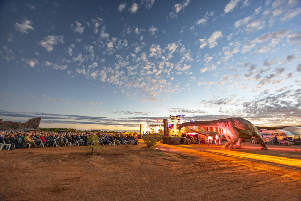 An outdoor event at sunset features a seated audience, a stage with lighting, and a large dinosaur sculpture in the foreground.