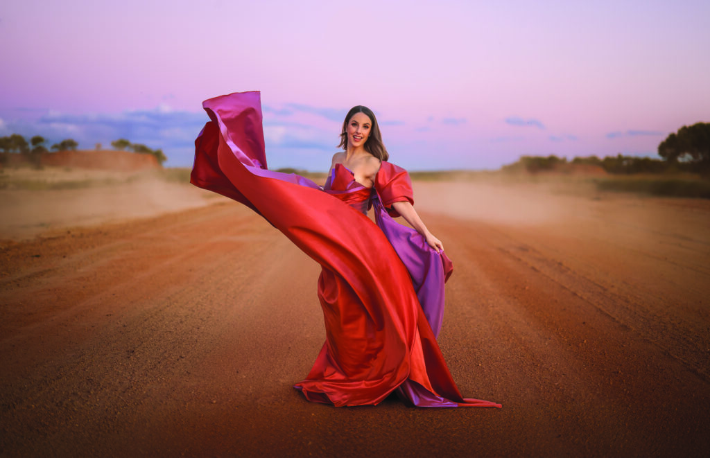 A woman in a flowing red and purple gown stands on a dirt road at sunset, with fabric billowing dramatically in the air around her.