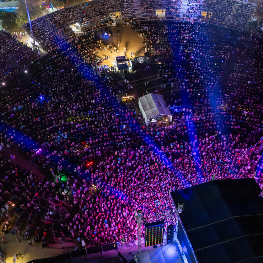 A large crowd fills an outdoor arena at night, illuminated by stage lights and blue beams, with a stage and equipment visible in the foreground.
