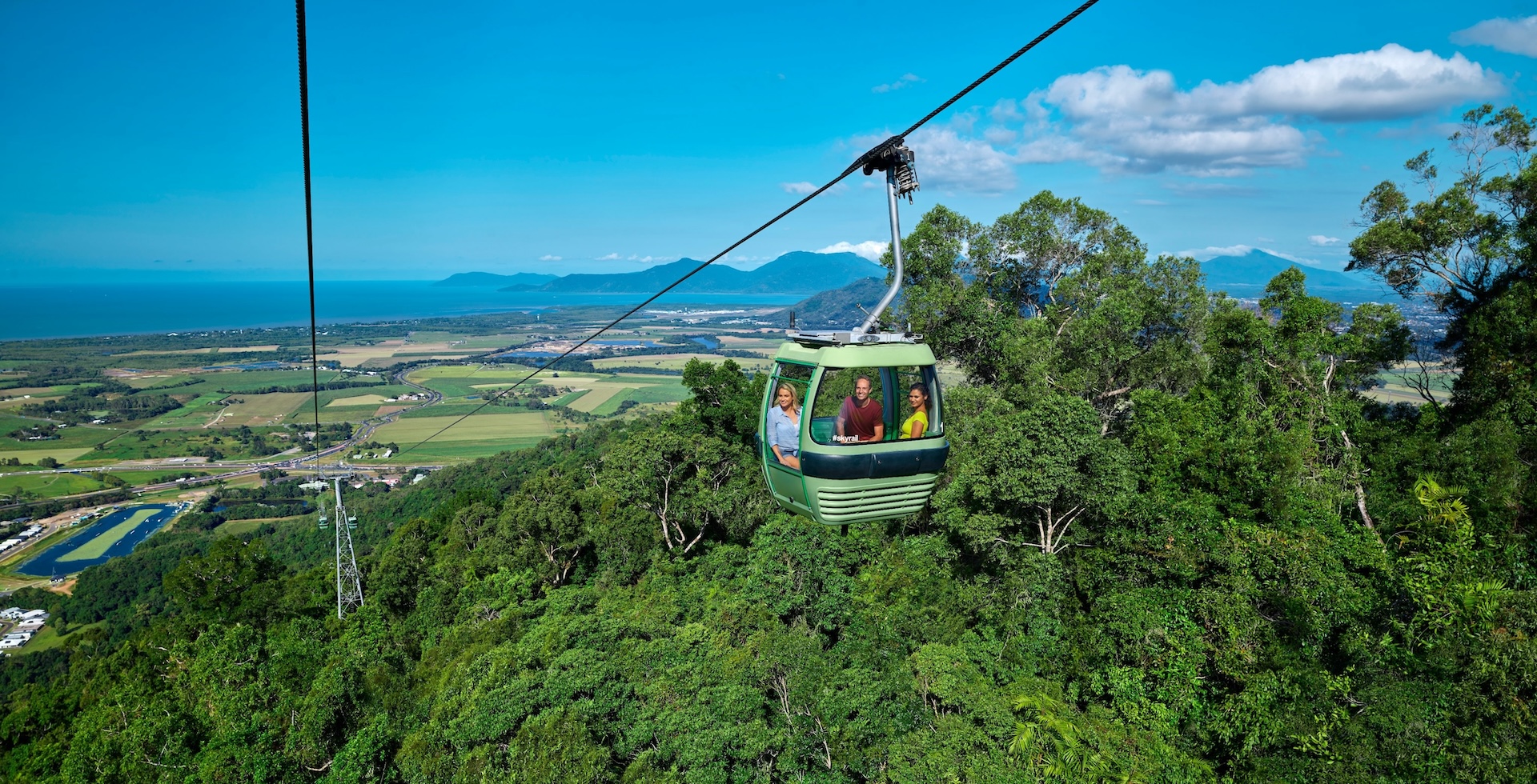 A green cable car carrying people travels over dense forest, with views of fields, coastline, and mountains under a bright blue sky.