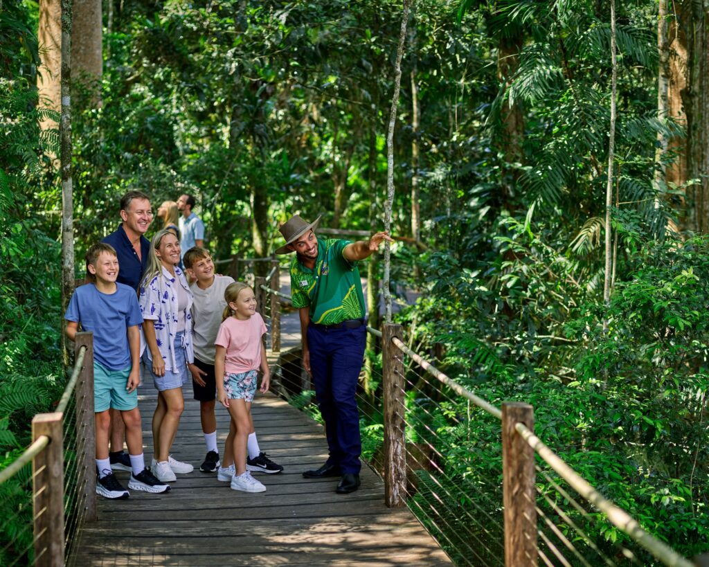 A group of people, including children and adults, stand on a wooden walkway in a lush forest as a guide points toward something in the trees.