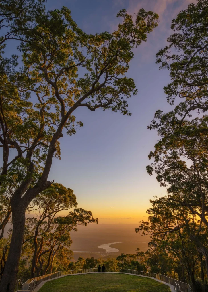 Two people stand at an overlook surrounded by tall trees, gazing at a river winding through a valley under a sunset sky.