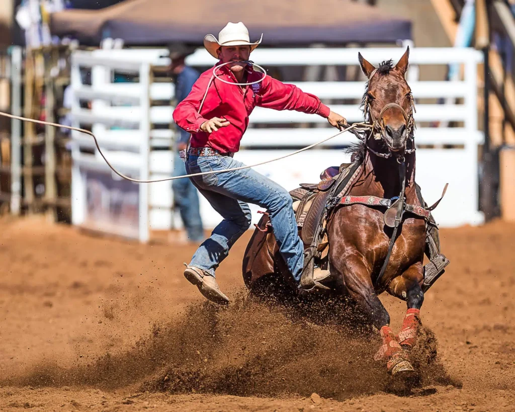 A cowboy in a red shirt and white hat leaps from his brown horse, holding a lasso, during a rodeo event on a dirt arena.