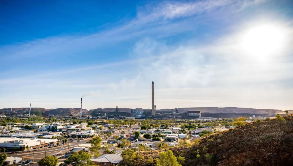 A panoramic view of a mining town under a bright sky with smokestacks emitting smoke in the background, surrounded by small buildings and trees.