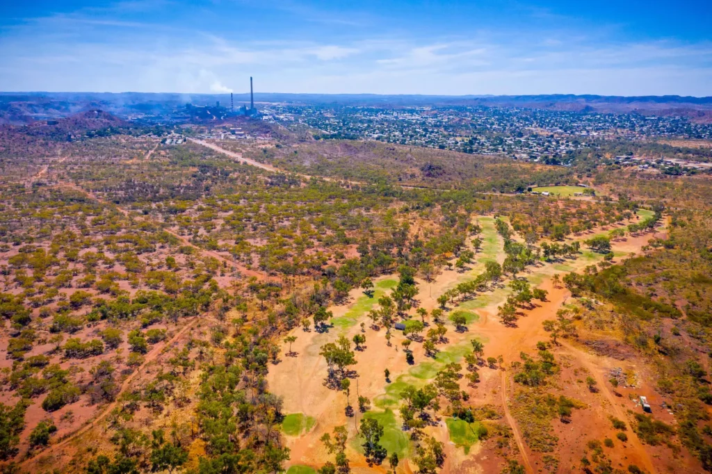 Aerial view of a sparsely vegetated landscape with a golf course. A town with industrial structures is in the background under a clear blue sky.