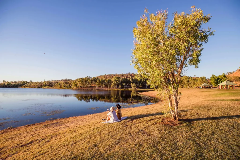 Two people sit by a tree on a grass field near the edge of a calm lake with clear blue skies and distant hills in the background.