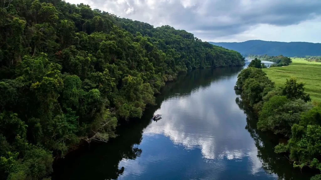 Dense green forest flanks a calm river reflecting clouds and sky, with a small boat visible on the water. Rolling hills are seen in the background under a partly cloudy sky.