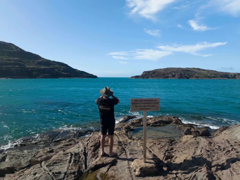 Person standing on a rocky shore, facing the ocean and taking photos with a camera. A nearby sign reads: 