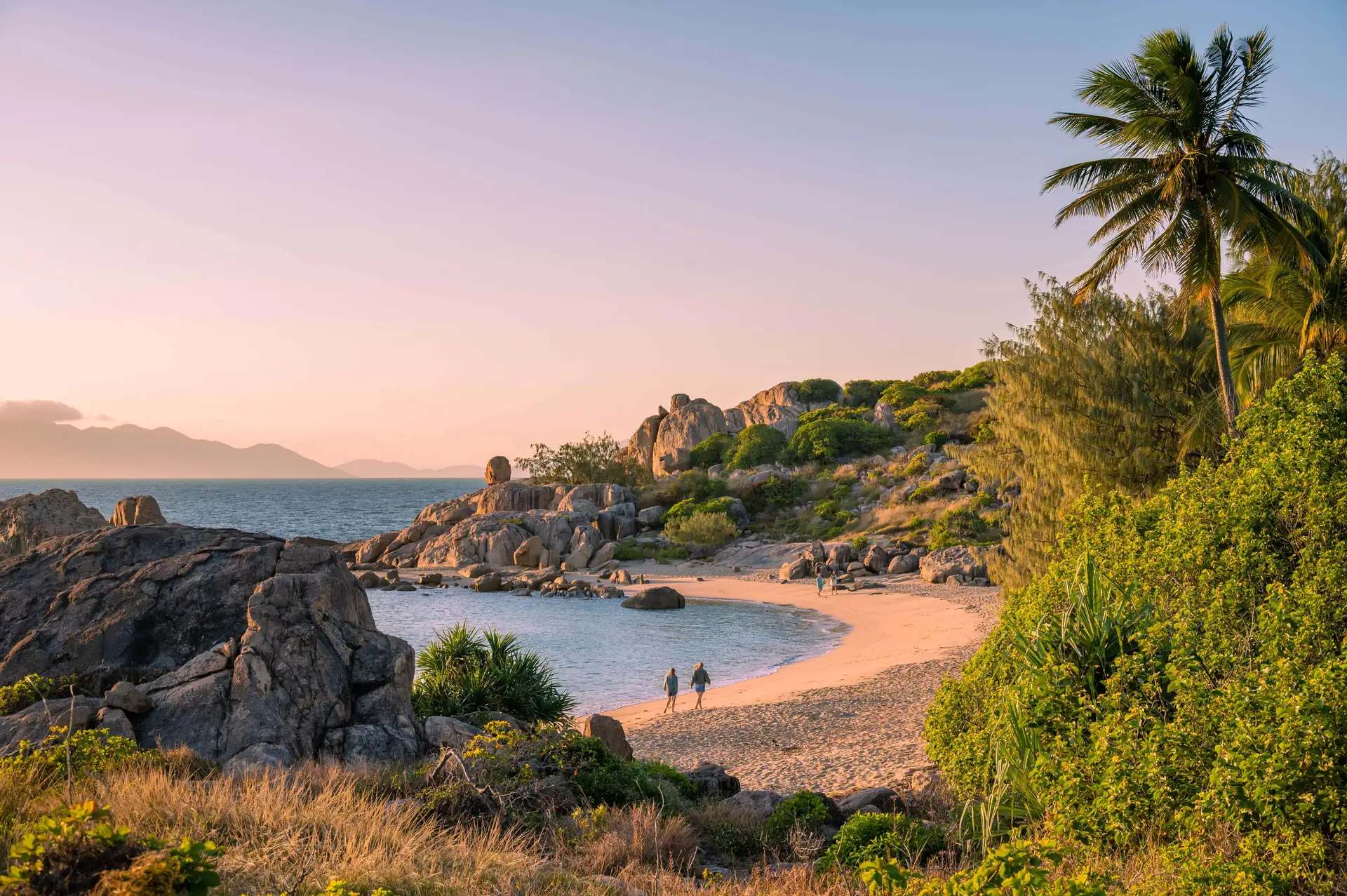 Two individuals walk along a secluded, rocky beach with a calm sea, lush vegetation, and distant mountains under a pink sky.