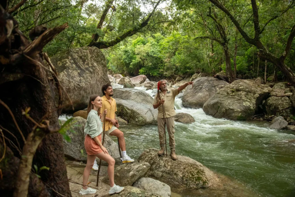 Three people, including a guide, standing on rocks by a river in a forest. The guide is pointing towards something in the distance.