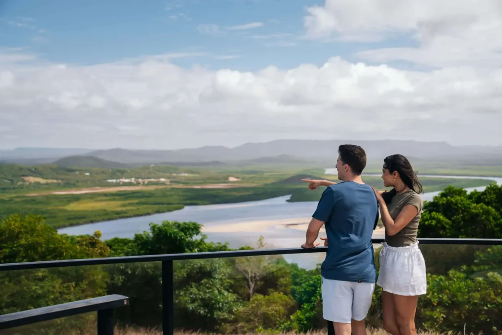 A man and woman stand on a viewing platform, looking at a scenic landscape with a river, green hills, and mountains in the distance. The man points towards the view.