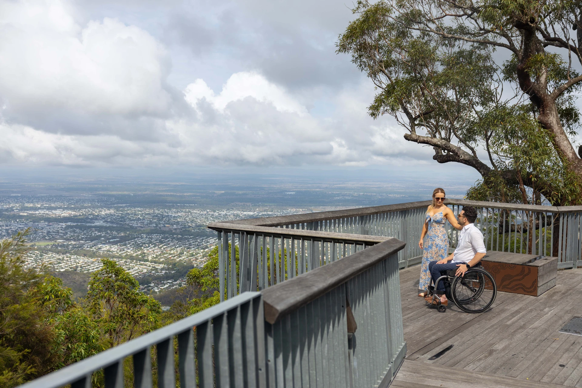 A woman and a man in a wheelchair on a wooden deck overlook a vast, cloudy landscape and city below.