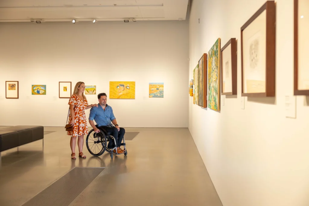 A woman and a man in a wheelchair are viewing paintings in an art gallery. The gallery features various colorful works on the walls.