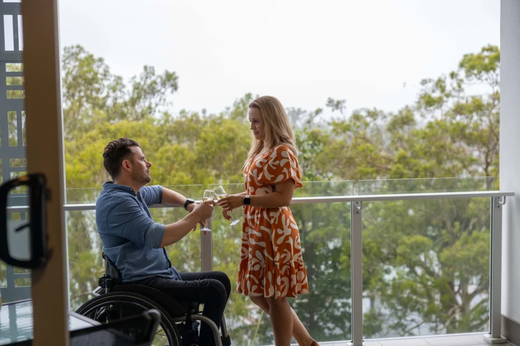 A man in a wheelchair and a woman in a patterned dress are standing and holding glasses on a balcony with a view of trees in the background.