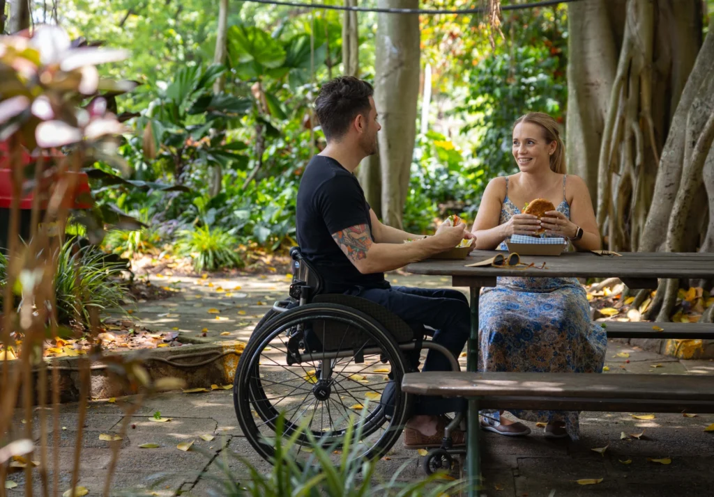 Two people having a meal at a picnic table in a lush garden. One person is in a wheelchair, and both are smiling and engaged in conversation.