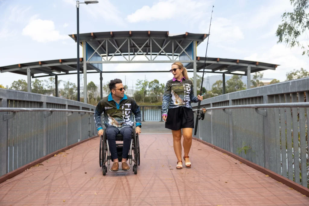 A man in a wheelchair and a woman holding a fishing rod walk on a bridge. Both are wearing fishing shirts and sunglasses. Trees and a covered structure are visible in the background.