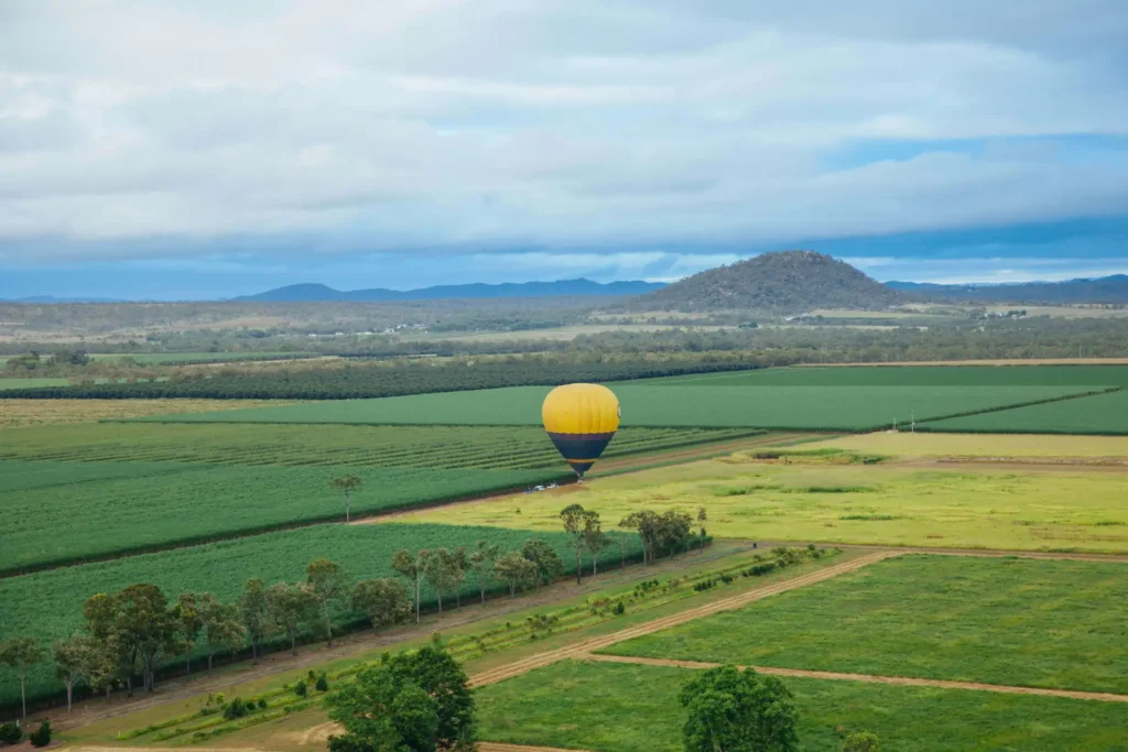 A yellow and black hot air balloon floats above a vast, green landscape with fields and distant hills under a partly cloudy sky.