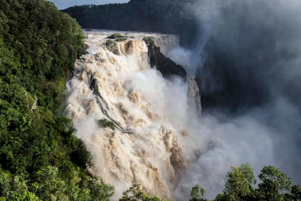 A powerful waterfall cascades down a cliff, surrounded by dense green forest, with mist rising from the intense water flow.