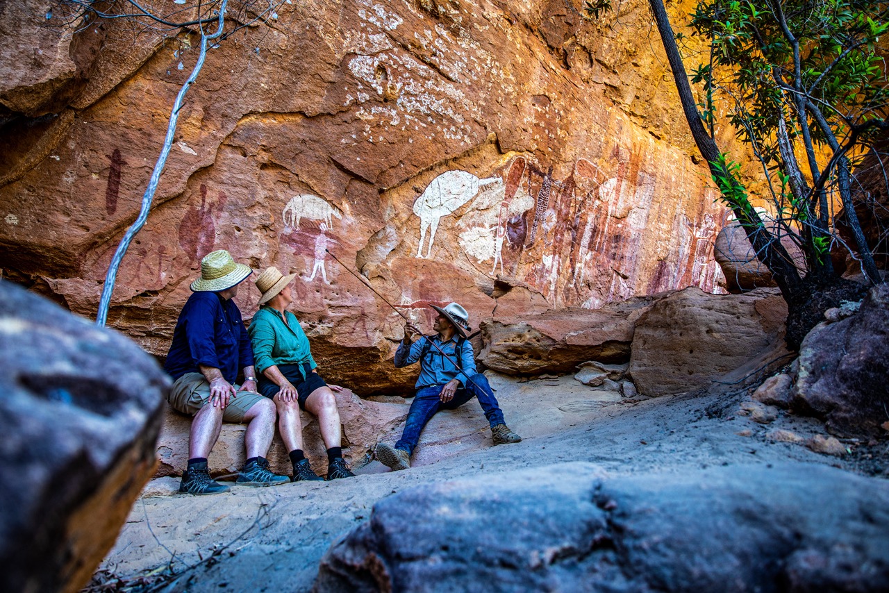 Three people sit on rocks by a cliff with ancient rock paintings in the background. They wear hiking attire and sun hats, engaging in conversation. The surrounding area is rocky with sparse vegetation.