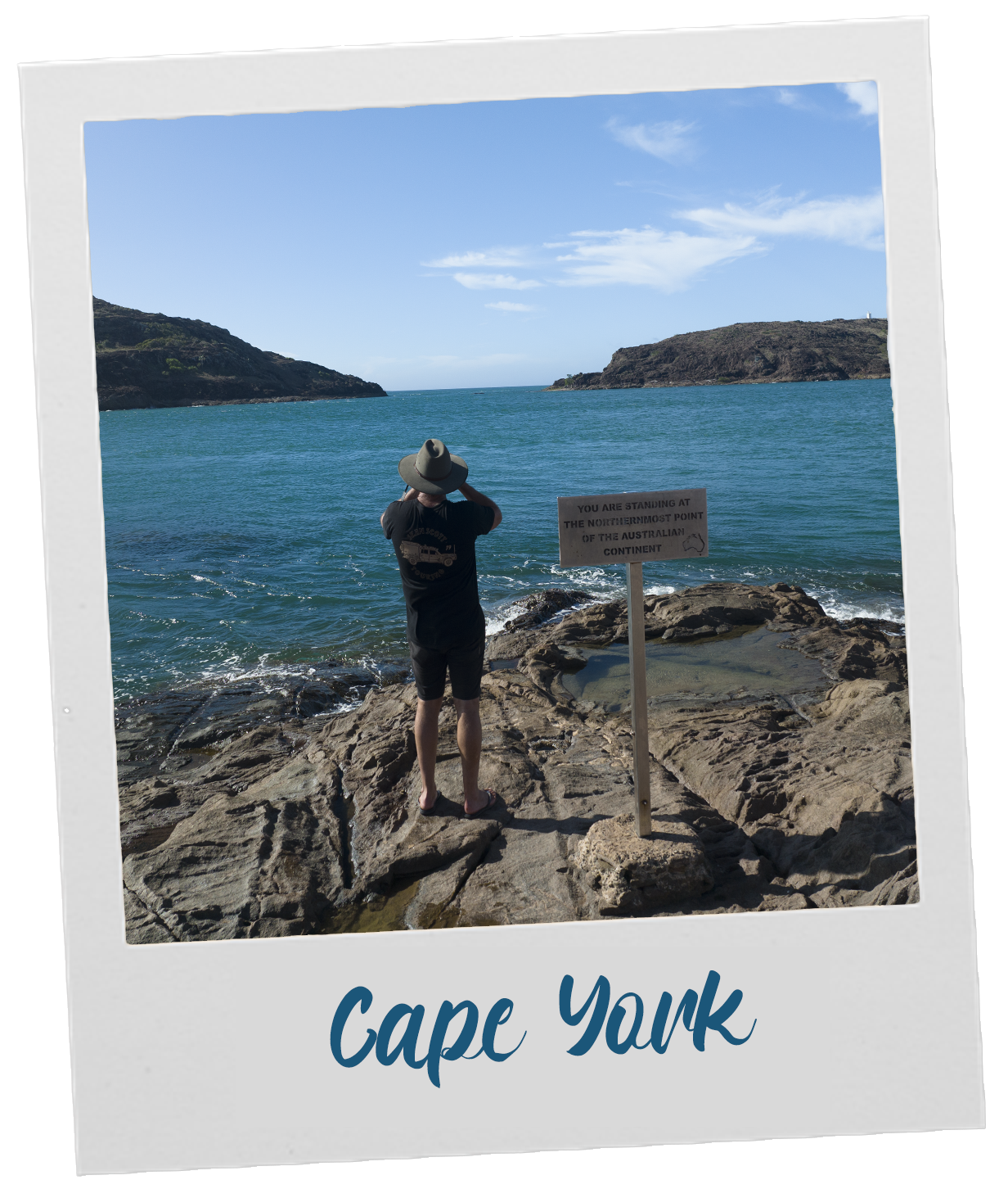 Person standing on rocky shore at Cape York, Australia, overlooking the ocean and taking a photo. A sign nearby reads 