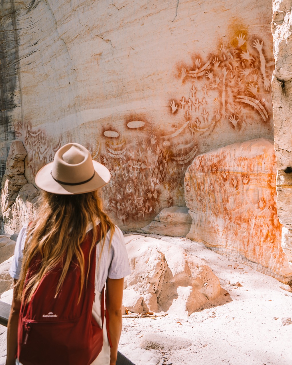 Person wearing a hat and red backpack looks at ancient rock art featuring handprints and faces on a sandstone wall.