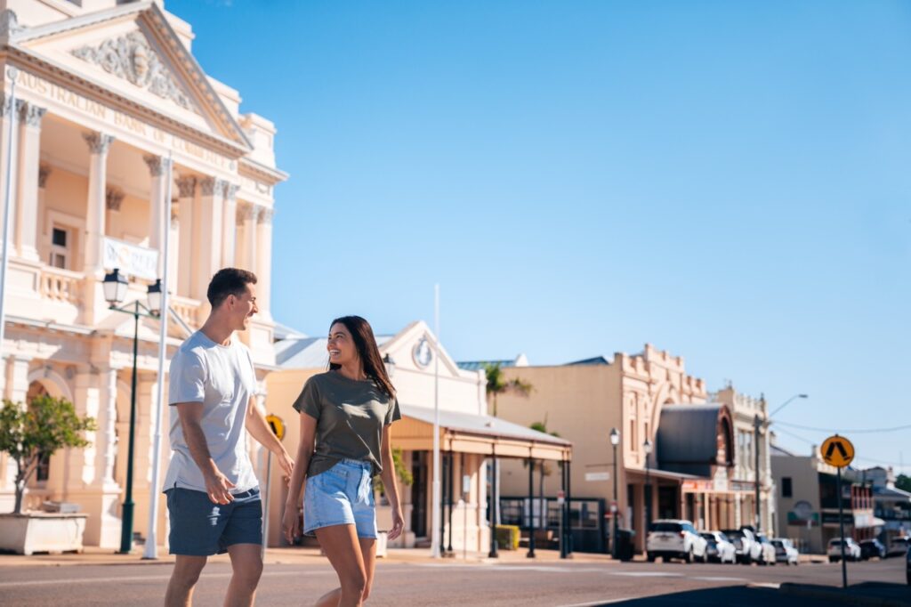 A man and woman walk hand in hand on a sunny day in front of a historical building on a street with old-style architecture.