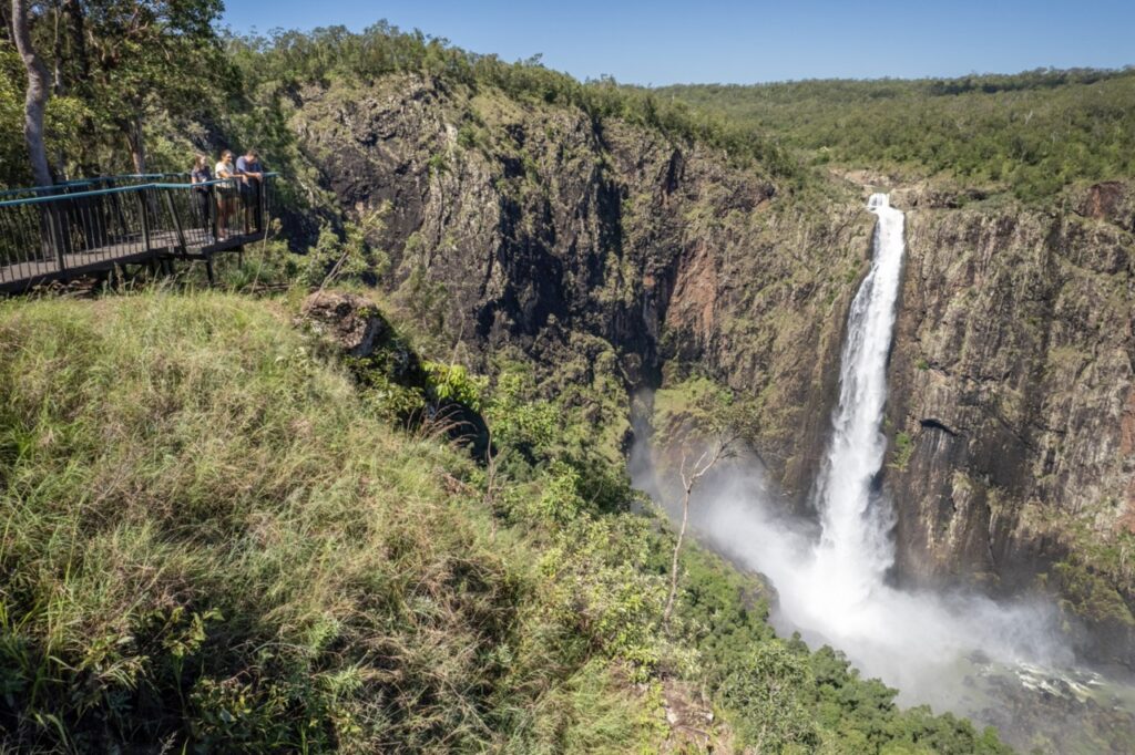 People stand on a viewing platform overlooking a large waterfall cascading down into a lush green valley with steep rocky cliffs.