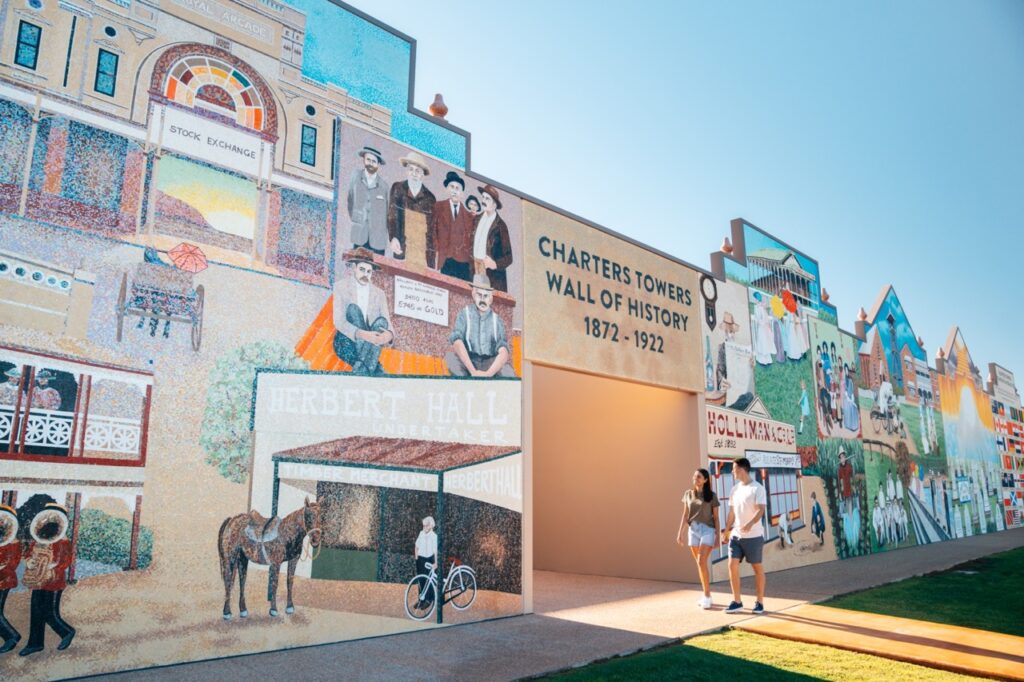 A couple walks past a historical mural depicting the history of Charters Towers from 1877 to 1922, featuring various scenes and structures, including Herbert Hall and horse-drawn carriages.