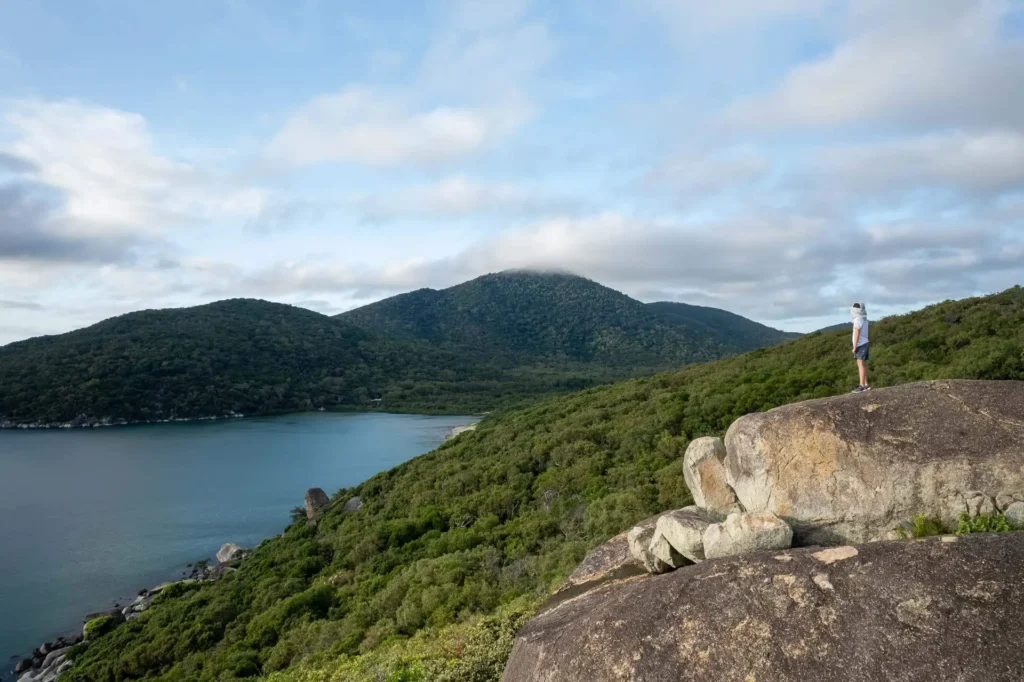 A person stands on a large rock overlooking a lush green landscape with a lake and mountains in the background under a partly cloudy sky.