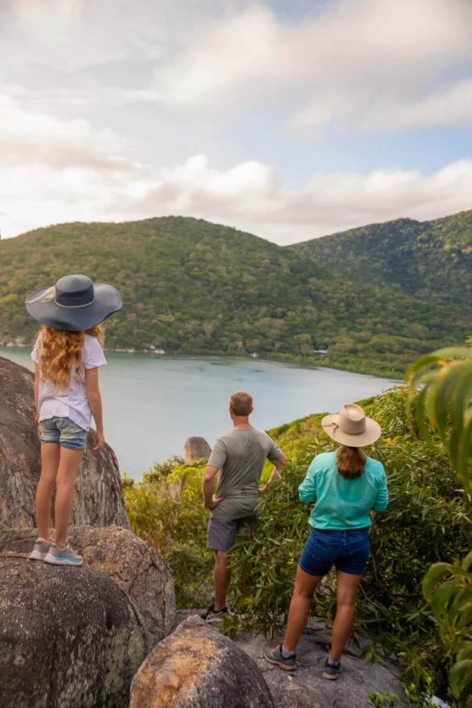 Three people in casual attire stand on rocks and look out at a lake surrounded by green hills, under a cloudy sky.