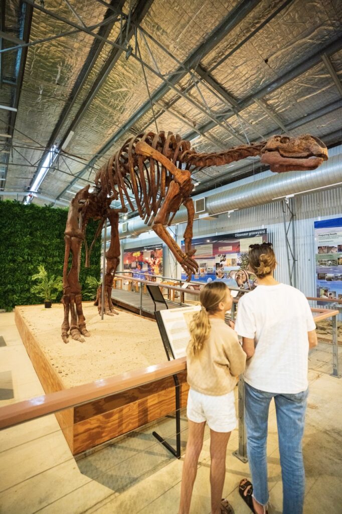 Two people observe a large dinosaur skeleton on display inside a museum exhibit hall.