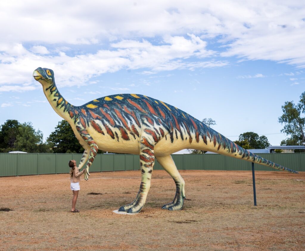 A person stands next to a large dinosaur statue with blue, orange, and yellow markings, situated in a fenced outdoor area.