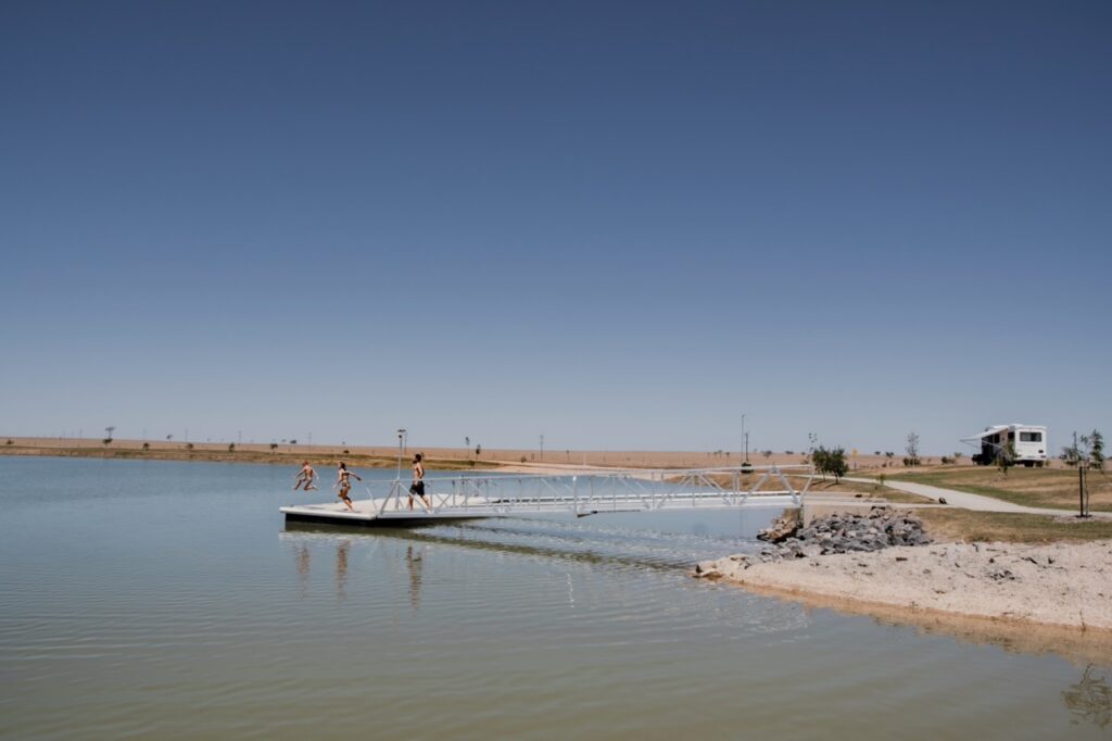 A few people are jumping off a narrow metal dock into a calm body of water under a clear blue sky. A house and trees are visible in the background.