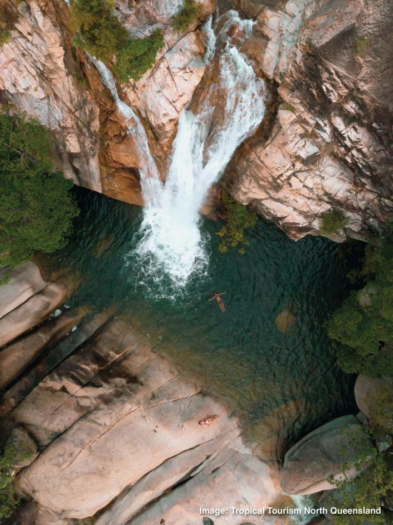 Aerial view of people swimming in a natural pool below a cascading waterfall surrounded by rocky cliffs and greenery. Image: Tropical Tourism North Queensland.