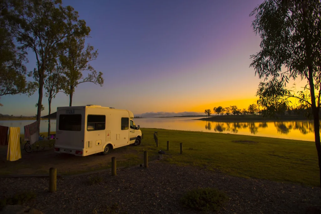 A white RV is parked by a lakeside at sunset, surrounded by trees and grassy land. The sky is a gradient of purple and orange, reflecting on the calm water.