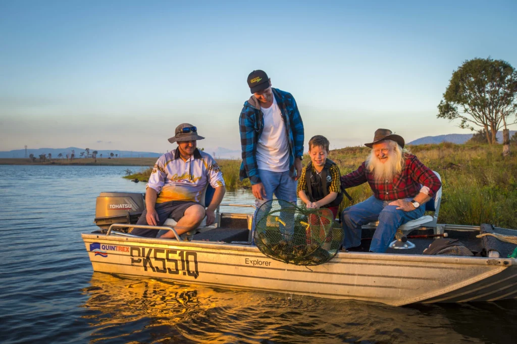 Three men and a boy sit in a small fishing boat on a calm lake at sunset. The boy holds a cage with fish while the others look on and smile. The boat has the number 