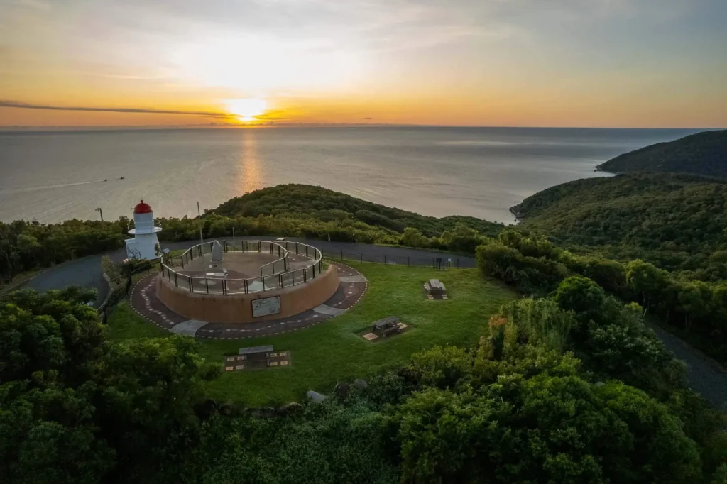 Aerial view of a coastal landscape featuring a white lighthouse and surrounding greenery with the sun setting over the ocean in the background.