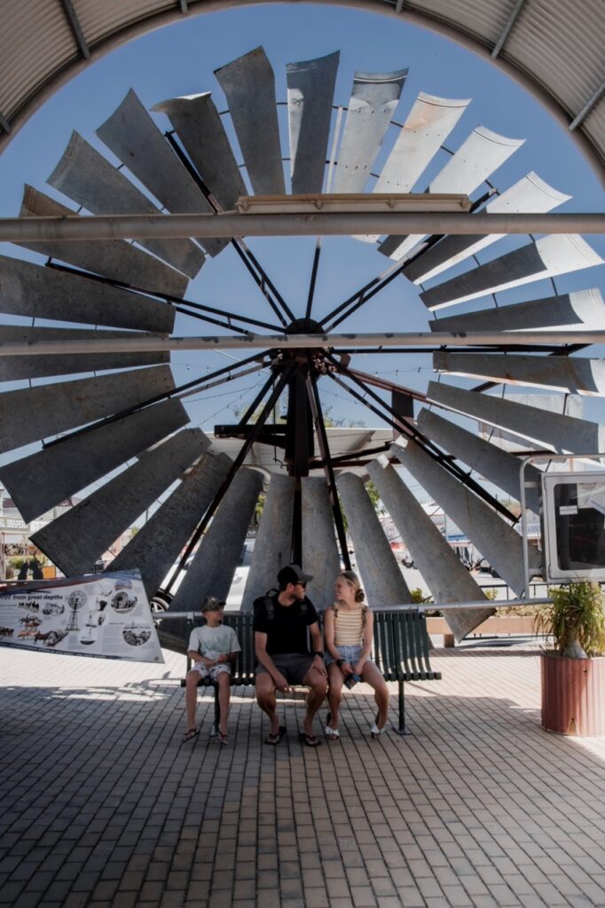 A group of three people sit on a bench under a large, metal windmill structure with blue skies in the background.