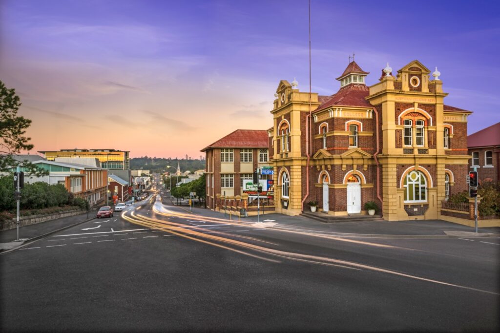 A historic building with ornate architecture is seen at a street intersection during sunset, with light trails from moving vehicles creating streaks on the road.