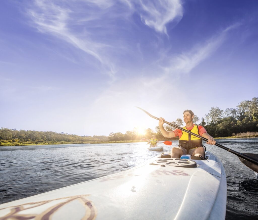 A person is paddling a kayak on a body of water, wearing a life jacket, with another kayaker visible in the distance under a clear blue sky.