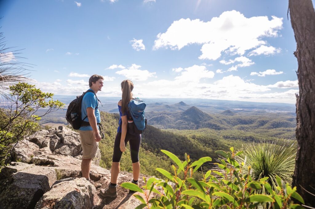 Two hikers with backpacks stand on a rocky cliff, overlooking a vast, forested landscape under a partly cloudy sky.