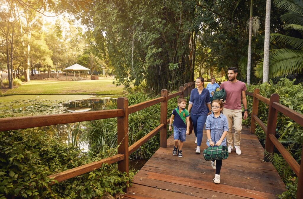 A family of five walks on a wooden bridge in a park, carrying picnic supplies. Trees and a pond are visible in the background.