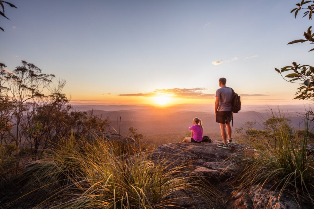 Two people are on a rocky cliff, one standing and one sitting, watching the sunset over a scenic landscape with mountains and trees.