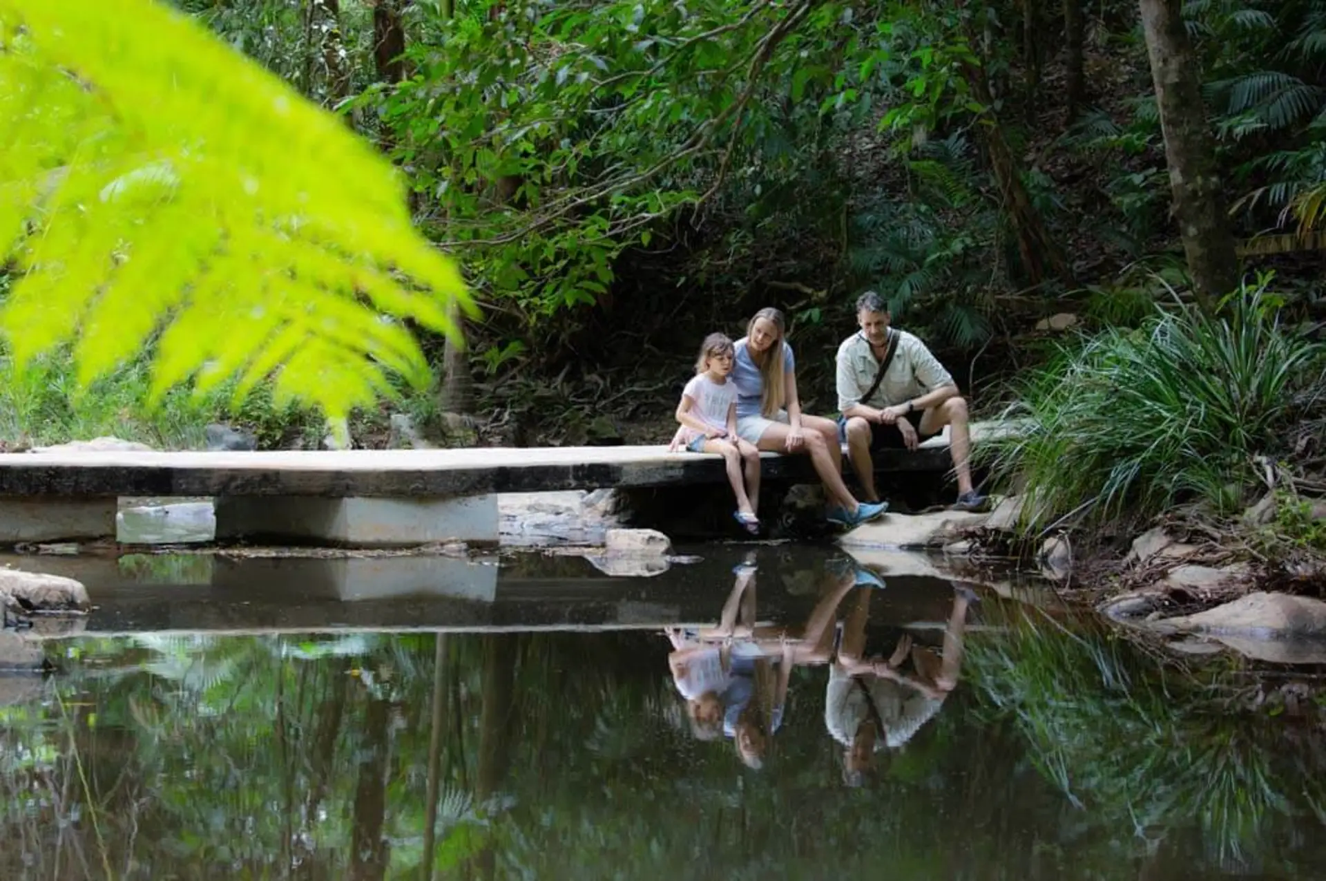 A family of three sits by a still pond in a forested area, their reflections visible in the water.