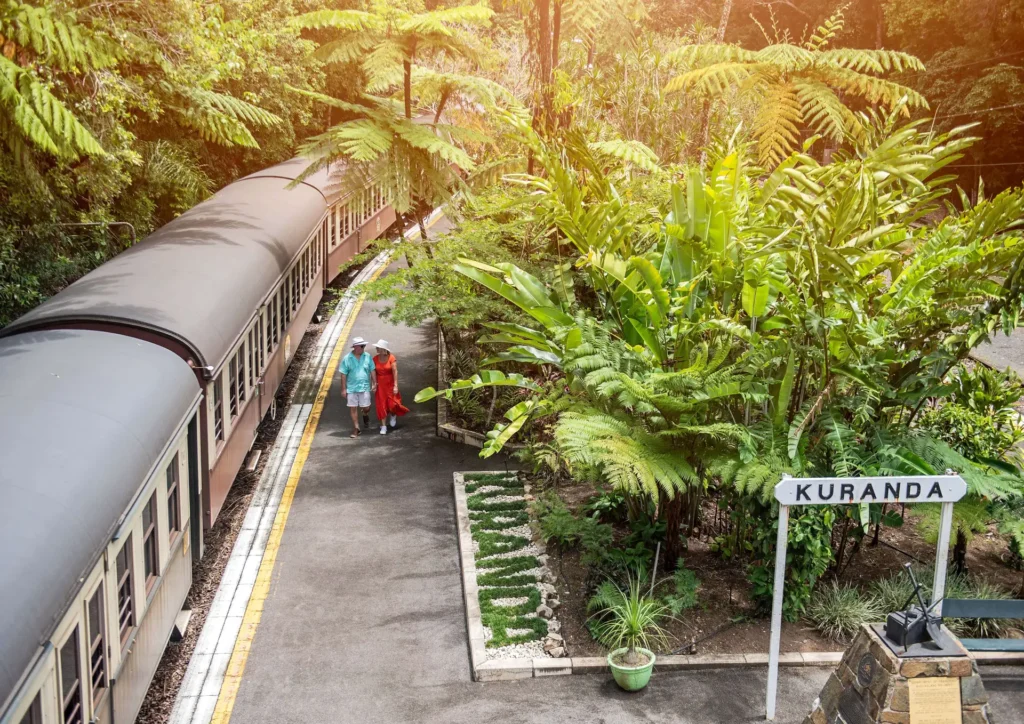 A train is at Kuranda station surrounded by tropical vegetation. A couple stands on the platform wearing casual clothes and hats.