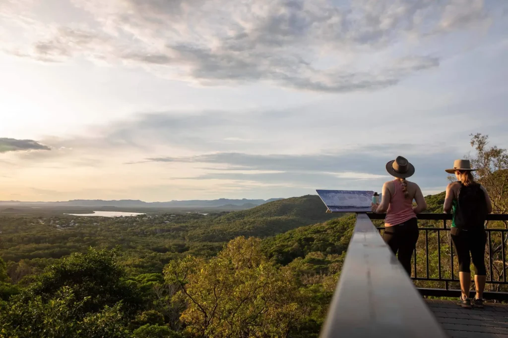 Two people stand on a viewing platform overlooking a lush, green landscape with hills and a distant body of water under a partly cloudy sky.