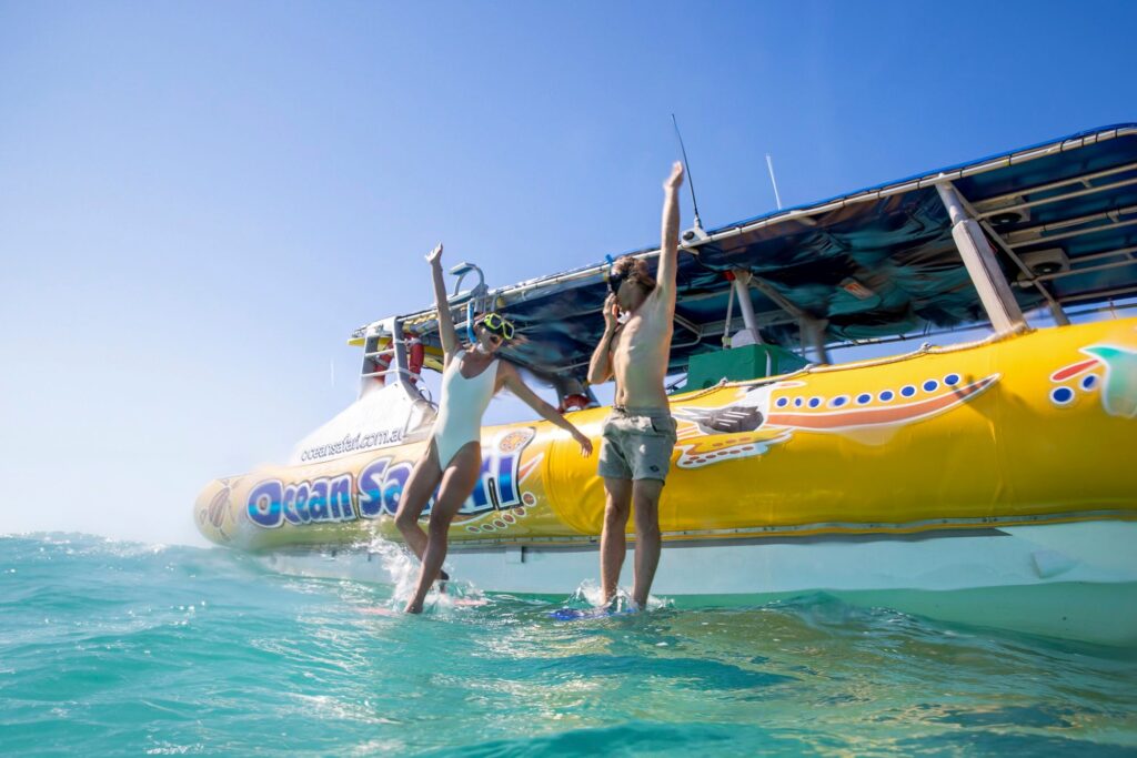 Two people in swimwear and snorkel masks jump off a yellow boat labeled 