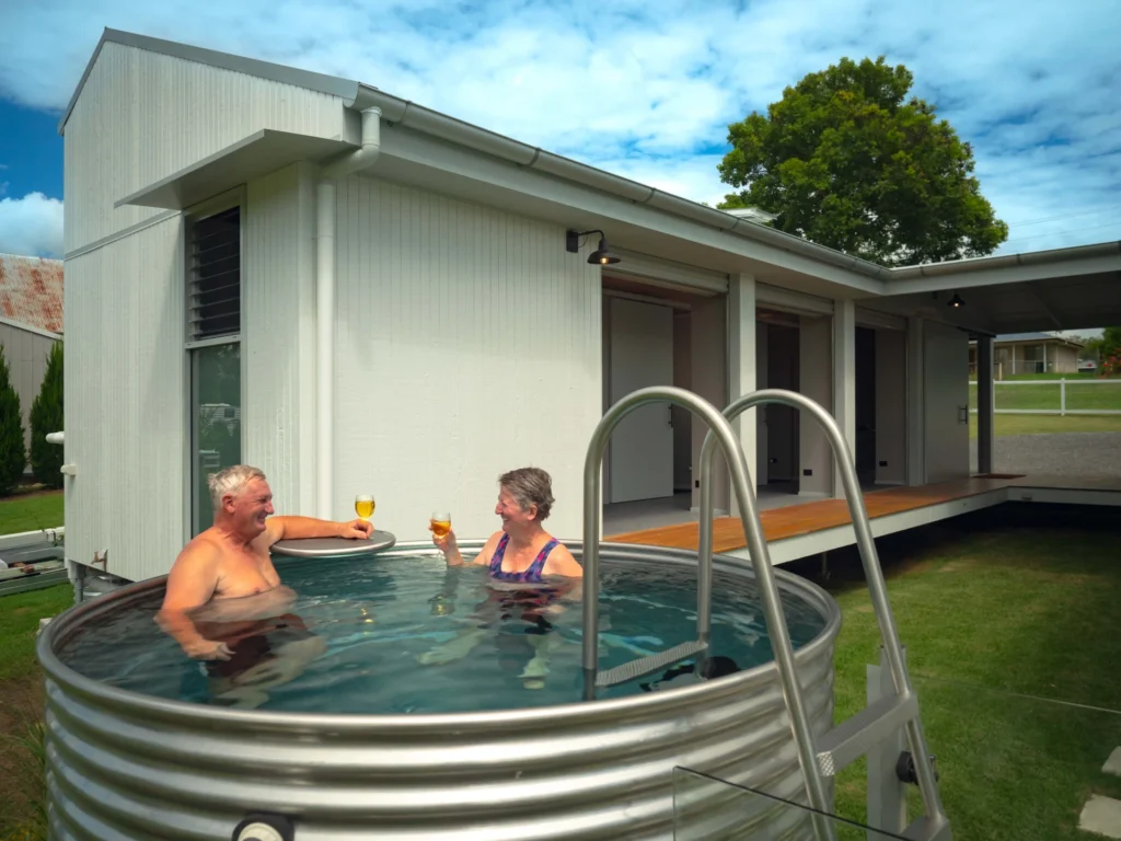 Two people relax in a galvanized steel hot tub, each holding a drink, outside a modern white house with a covered patio and trees in the background.