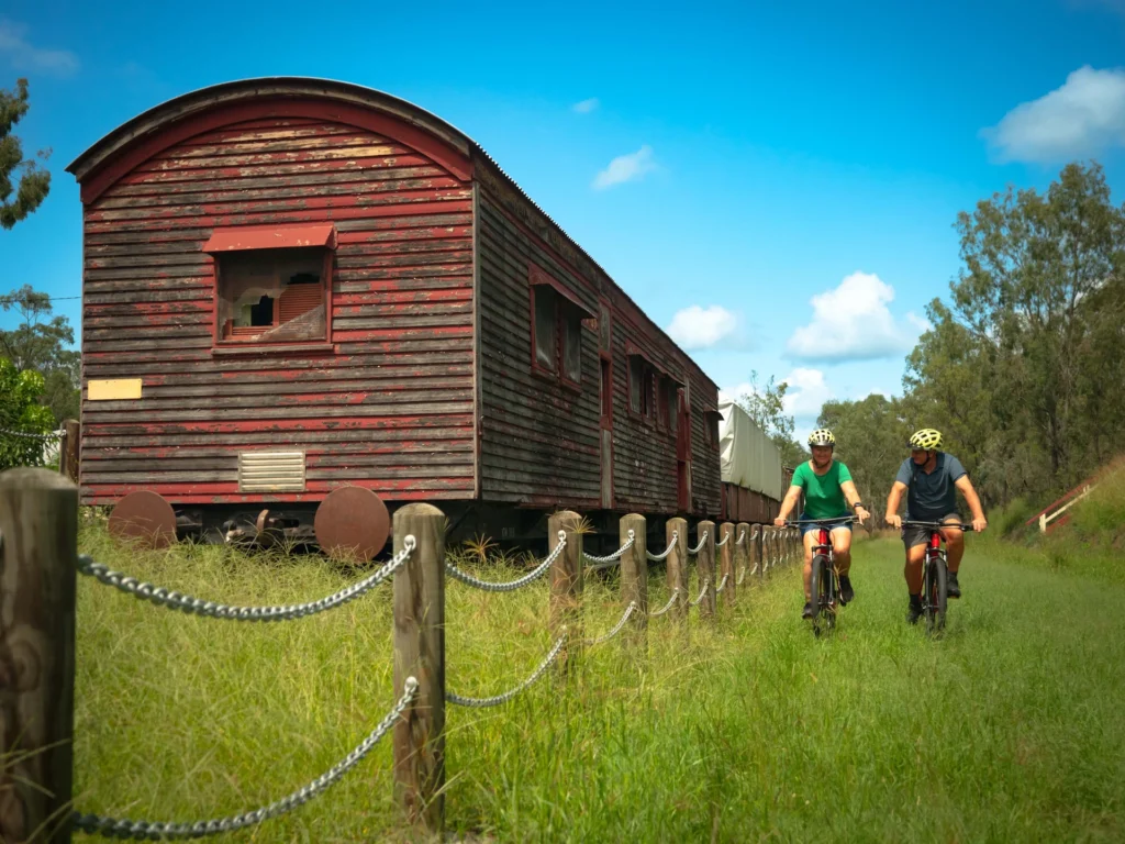 Two people ride bicycles along a grassy path beside an old, red, weathered train car under a clear blue sky.