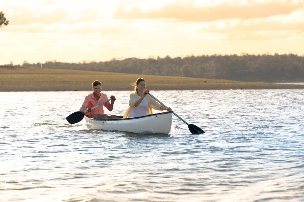 Two people paddle a canoe on a calm body of water with distant trees and an evening sky in the background.